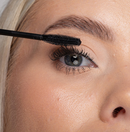 Žena si pečlivě nanáší řasenku na řasy pomocí kartáčku na řasenky, čímž přesně zvýrazňuje její oční make-up.